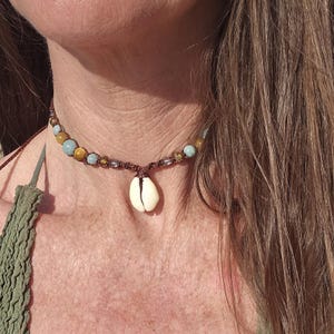 Puede incluir: Un collar tipo gargantilla con un colgante central de concha de cauri blanca suspendido de un cordón marrón. El cordón está adornado con una serie de pequeñas cuentas redondas y multicolores en tonos azul claro, ámbar y marrón translúcido.