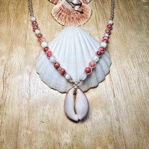 May include: A necklace featuring a central white cowrie shell pendant suspended from a tan cord. The cord is adorned with alternating round beads in shades of mottled pink and smooth white, creating a symmetrical pattern along the neckline.