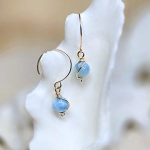 Puede incluir: Un par de pendientes colgantes con cuentas esféricas de color azul claro envueltas en alambre dorado. Los pendientes tienen un diseño simple y elegante con un gancho dorado. Los pendientes se exhiben sobre una superficie blanca texturizada.