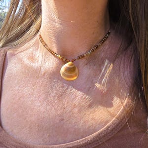 May include: A seashell pendant necklace with a brown beaded chain. The pendant is a light tan color with brown stripes. The necklace is worn around the neck and is a natural, earthy accessory.