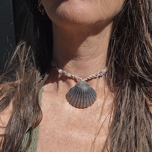 May include: A choker necklace featuring a central dark grey, ribbed scallop shell pendant suspended from a woven beige cord. The cord is accented with small, multi-colored round beads in shades of white, pink, and grey along the neckline.