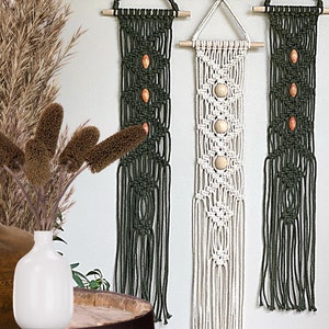 May include: Three macrame wall hangings with wooden beads. The hangings are in different colors: green, white, and beige. The hangings are all the same size and shape.