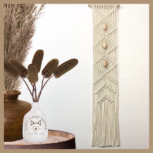May include: A beige macrame wall hanging with a geometric pattern and three wooden beads. The hanging is displayed on a white wall with a vase of brown dried flowers in the foreground.
