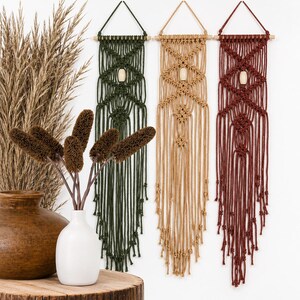 Puede incluir: Tres tapices de macramé en verde, beige y burdeos, cada uno con una clavija de madera y una cuenta central de madera. Los tapices se exhiben contra una pared blanca, con un jarrón de flores secas y un jarrón decorativo sobre una superficie de madera.