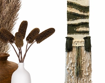 May include: A woven wall hanging with a wooden dowel at the top. The tapestry features a design of olive green and cream-colored horizontal stripes and textured sections. The bottom has long, cream-colored fringe and olive green tassels.