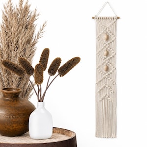 May include: A long, vertical macrame wall hanging made of off-white cotton cord, featuring a diamond knot pattern with three smooth, light-colored wooden beads integrated into the center, suspended from a horizontal wooden dowel with a white cord.