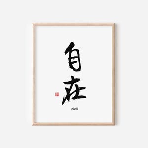 May include: A framed print featuring black calligraphy on a white background. The artwork displays two Chinese characters and the English translation "zi zai" at the bottom. A small red stamp is present near the bottom. The frame is a light wood color.