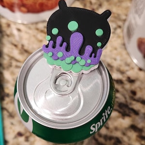 May include: A black, purple, and light green 3D-printed plastic soda can tab opener shaped like a stylized creature with a dripping liquid design. The accessory is attached to the silver pull tab of a green Sprite lemon-lime soda aluminum can.