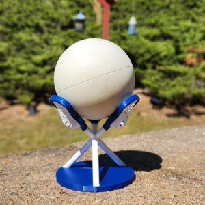 May include: A white sphere rests on a blue and white stand. The stand has a circular base and supports the sphere with a crisscross design. The stand is on a concrete surface, with a blurred green background.