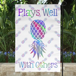 May include: A white garden flag with a rainbow pineapple design and the text "Plays Well With Others".