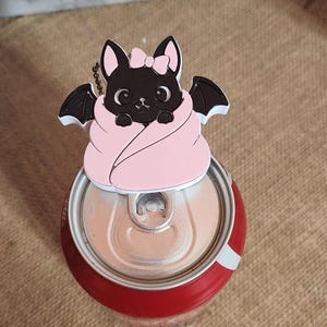 May include: A black cat-shaped beverage can tab opener featuring a light pink blanket wrap, a small pink bow on its head, and black bat wings. The accessory is positioned on the top of a red aluminium soda can with a silver pull tab mechanism.
