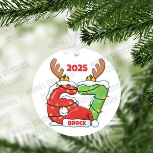 May include: A white Christmas ornament with a festive design. The ornament features the numbers 6 and 7, decorated with Christmas lights and reindeer antlers, and the year 2025. The name "BROCK" is displayed on a red hat.