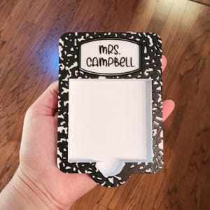 May include: A black and white composition notebook-themed holder with the text "Mrs. Campbell" in a white rectangle. The holder has a white interior and is held in a hand.