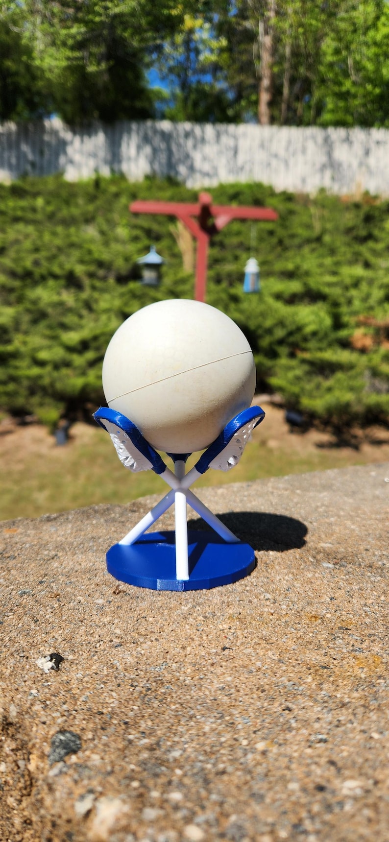 May include: A white sphere rests on a blue and white stand. The stand has a circular base and supports the sphere with a crisscross design. The stand is on a concrete surface, with a blurred green background.