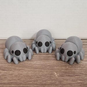 Puede incluir: Tres figuras de arañas grises impresas en 3D con ojos negros y un cuerpo segmentado. Las arañas se exhiben sobre una superficie de madera sobre un fondo blanco. Las figuras son un artículo decorativo caprichoso.