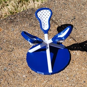 May include: A blue and white lacrosse stick-themed decorative object. The object has a blue circular base, white support beams, and a blue and white lacrosse stick head. The object is sitting on a textured surface.