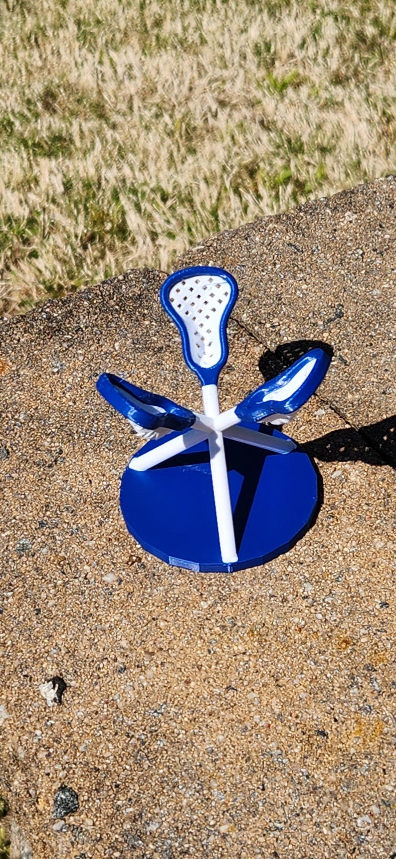 May include: A blue and white lacrosse stick-themed decorative object. The object has a blue circular base, white support beams, and a blue and white lacrosse stick head. The object is sitting on a textured surface.