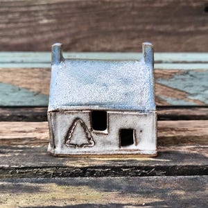 May include: A miniature ceramic house with a pale blue roof and white walls. The house features two small windows and a tree design. It is placed on a weathered wooden surface, evoking a rustic aesthetic.