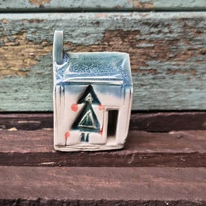 Könnte beinhalten: Eine kleine Keramikhausfigur mit einer glänzenden blau-weißen Glasur. Die Vorderseite zeigt ein dunkelgrünes, dreieckiges Baummotiv, kleine orangefarbene kreisförmige Akzente und einen rechteckigen Ausschnitt als Türöffnung.
