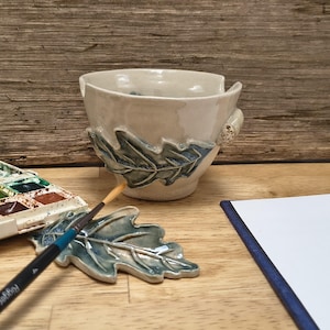 Può includere: Una ciotola in ceramica per filati con smalto lucido bianco sporco e un rilievo di foglia di quercia blu-verde scolpito sul lato. Una foglia di quercia in ceramica abbinata si trova nelle vicinanze, accompagnata da un pennello sottile e una tavolozza.