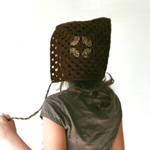 Può includere: Un cappello con cappuccio in crochet marrone con un motivo floreale in un colore marrone più chiaro. Il cappello è indossato da una persona con capelli lunghi e castani.