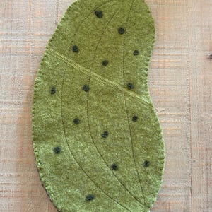 May include: A green, bean-shaped felt trivet with dark green accents. The trivet has a stitched design with dark green, circular embellishments. The edges are finished with a stitched border. The trivet is designed to protect surfaces from heat.