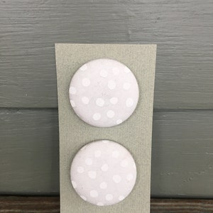 May include: Two round, light gray buttons with white polka dots are attached to a rectangular, light green card. The buttons are evenly spaced and centered on the card, which is set against a gray wooden background.