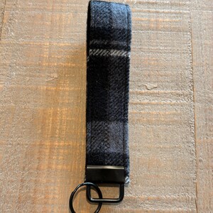 May include: A black and gray plaid wristlet keychain. The fabric wrist strap is attached to a black metal rectangle and a black metal split ring. The plaid pattern features shades of gray and black.