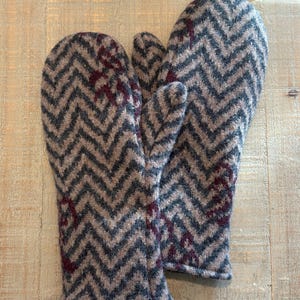 May include: A pair of knitted mittens with a chevron pattern in gray, blue, and brown. The mittens have a cuff and a thumb. There are maroon accents on the front of the mittens. The mittens are made of a soft, warm material.