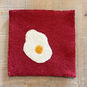May include: A square, red felt trivet with a white and yellow fried egg design. The egg is centered on the trivet and has a textured, hand-stitched appearance. The trivet's edges are neatly finished with a contrasting stitch.