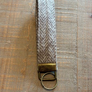 May include: A key fob with a herringbone pattern in shades of gray and brown. The fabric loop is attached to a bronze-colored metal clasp and key ring. The key fob is designed to hold keys.