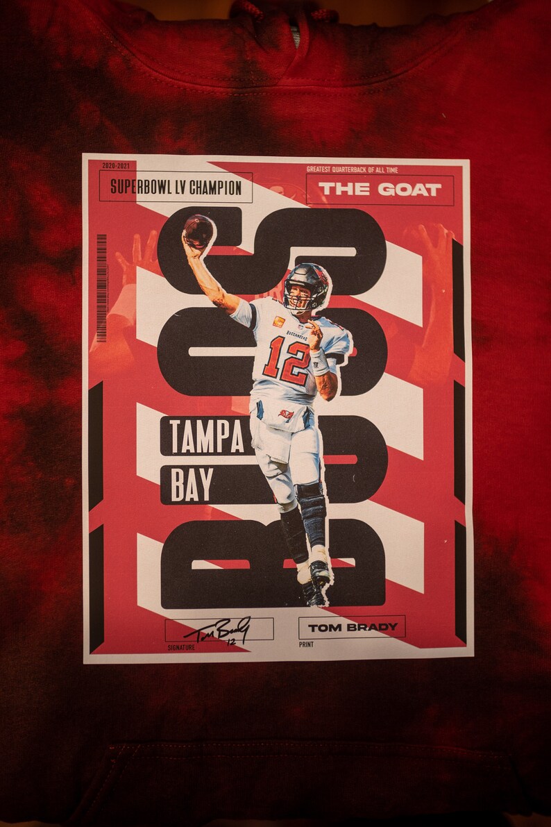 May include: A red and white graphic design with a photo of Tom Brady in a Tampa Bay Buccaneers uniform. The text "2020-2021 Superbowl LV Champion" and "The Goat" are included in the design.