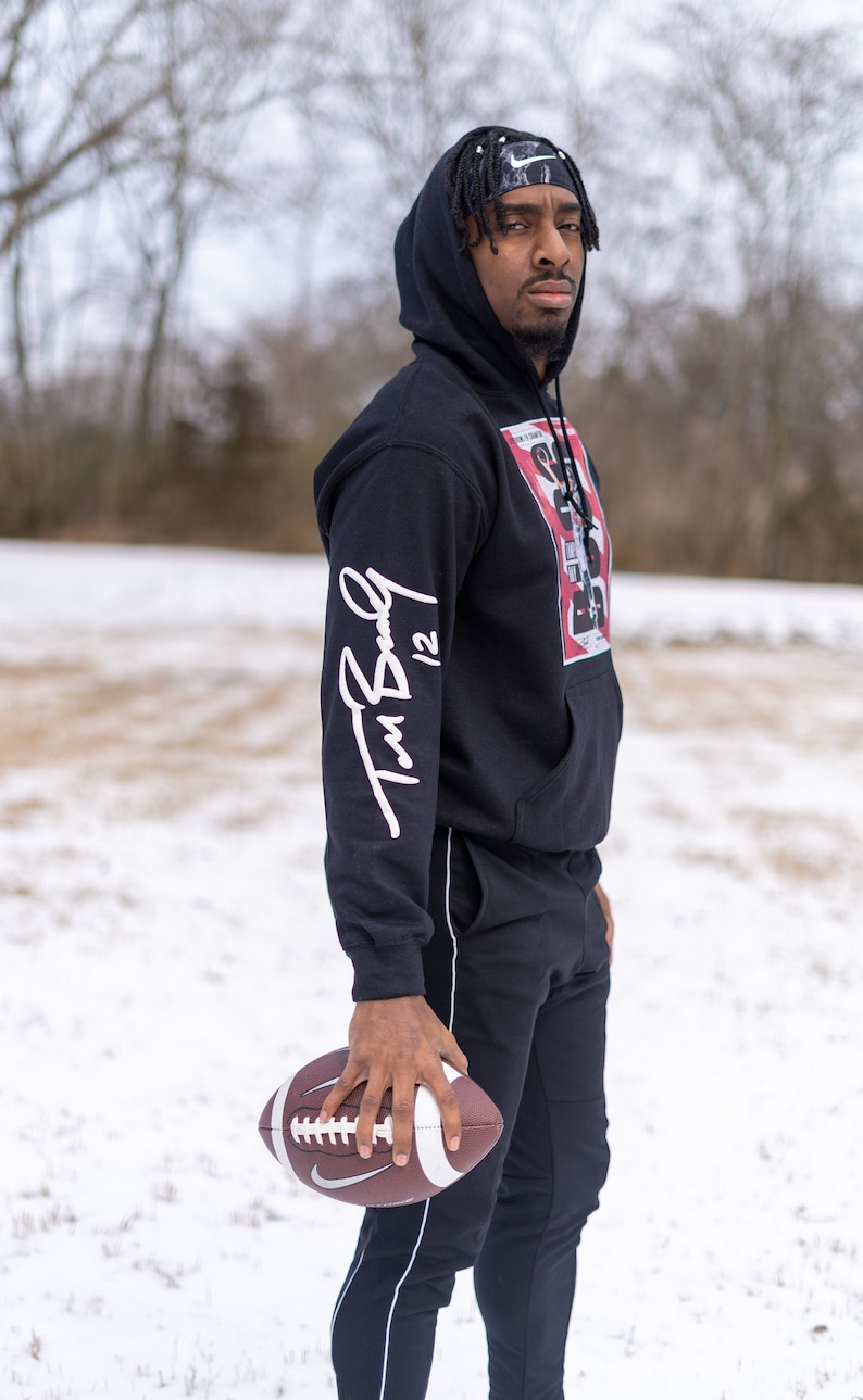 May include: A person wearing a black hoodie with a white signature on the sleeve and a graphic print on the front. The person is holding a brown and white football. The person is standing in a snowy field.