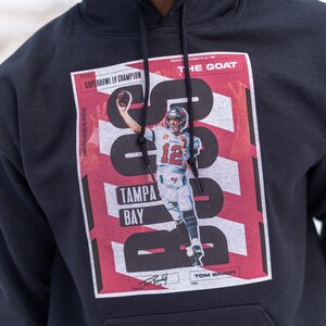May include: Black hoodie with a red and white graphic print featuring a football player in action. The graphic includes the text "Super Bowl LV Champion", "Tampa Bay", "The Goat", and "Tom Brady".