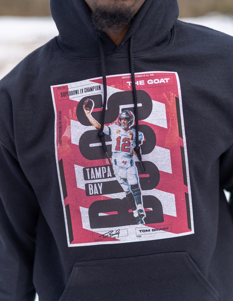 May include: Black hoodie with a red and white graphic print featuring a football player in action. The graphic includes the text "Super Bowl LV Champion", "Tampa Bay", "The Goat", and "Tom Brady".