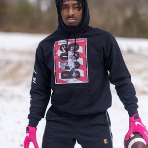 May include: A person wearing a black hoodie with a graphic of Tom Brady on the front. The graphic features the text "The Goat" and "Tampa Bay". The person is wearing pink gloves and holding a brown football.