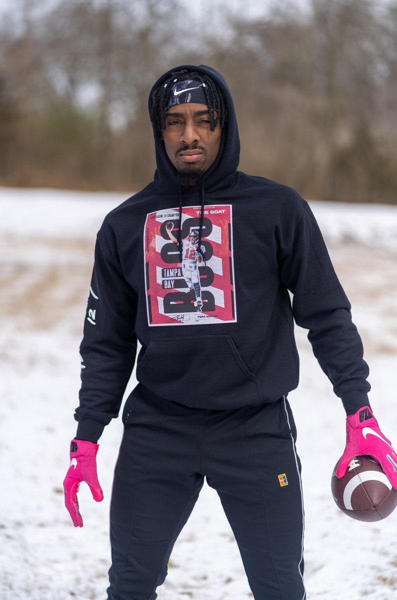 May include: A person wearing a black hoodie with a graphic of Tom Brady on the front. The graphic features the text "The Goat" and "Tampa Bay". The person is wearing pink gloves and holding a brown football.