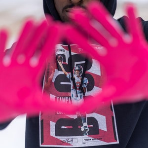 May include: A black hoodie with a red and white graphic print featuring Tom Brady, the text "Tampa Bay", and the number "12". The person is wearing pink gloves and making a heart shape with their hands.