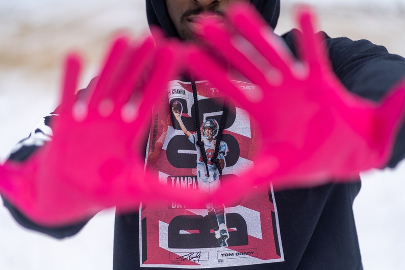 May include: A black hoodie with a red and white graphic print featuring Tom Brady, the text "Tampa Bay", and the number "12". The person is wearing pink gloves and making a heart shape with their hands.