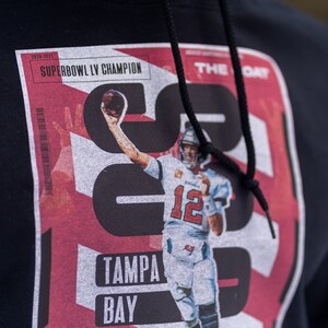 May include: Black hoodie with a red, white, and black graphic print of a football player holding a football. The graphic includes the text "SUPERBOWL LV CHAMPION", "TAMPA BAY", and "THE DAY".