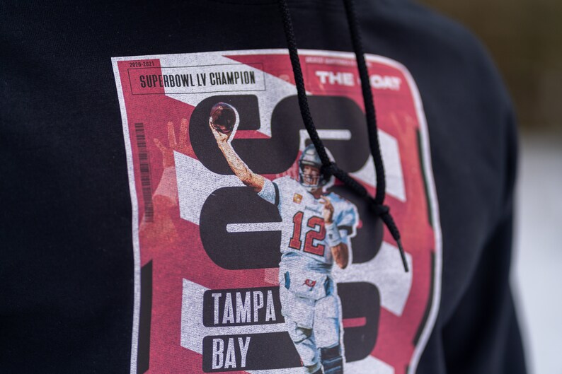 May include: Black hoodie with a red, white, and black graphic print of a football player holding a football. The graphic includes the text "SUPERBOWL LV CHAMPION", "TAMPA BAY", and "THE DAY".