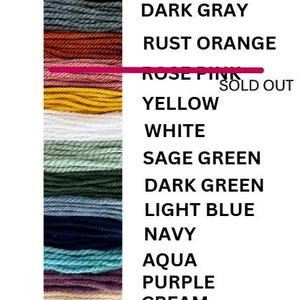 May include: A color chart featuring a vertical arrangement of yarn samples in various hues. The colors include black, dark gray, rust orange, yellow, white, sage green, dark green, light blue, navy, aqua, purple, cream, and brown. The text "ROSE PINK SOLD OUT" is also present.