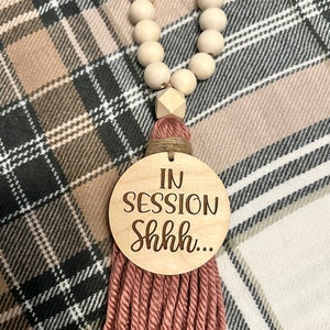 May include: Wooden bead garland with a decorative tassel and a round wooden sign that reads "IN SESSION Shhh...". The tassel is made of pink yarn and the beads are light brown.
