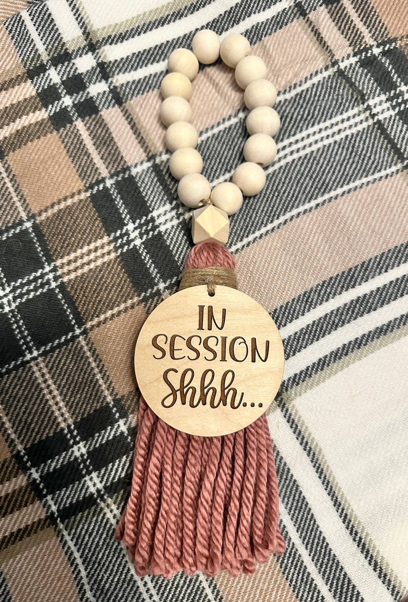 May include: Wooden bead garland with a decorative tassel and a round wooden sign that reads "IN SESSION Shhh...". The tassel is made of pink yarn and the beads are light brown.
