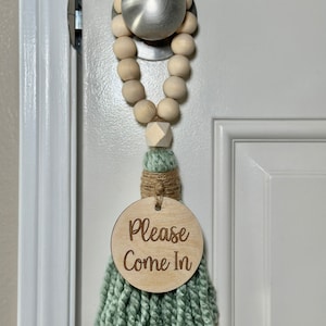 May include: A door hanger with a wooden bead garland and a mint green tassel. The round wooden sign reads "Please Come In". The door hanger is attached to a silver door knob.