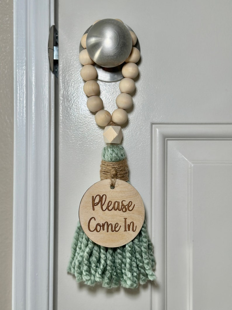 May include: A door hanger with a wooden bead garland and a mint green tassel. The round wooden sign reads "Please Come In". The door hanger is attached to a silver door knob.