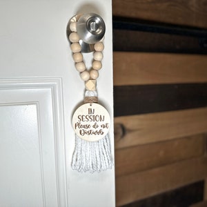 May include: A wooden door hanger with the text "IN SESSION Please do not Disturb" is suspended from a door handle. The hanger features a wooden bead garland, a white tassel, and a round wooden sign with engraved text.