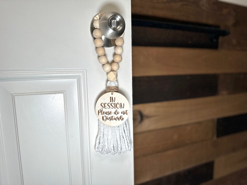 May include: A wooden door hanger with the text "IN SESSION Please do not Disturb" is suspended from a door handle. The hanger features a wooden bead garland, a white tassel, and a round wooden sign with engraved text.
