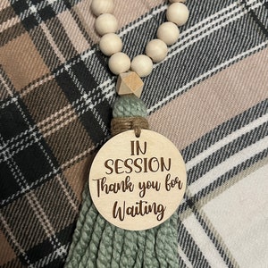 May include: A wooden bead garland with a sage green tassel and a round wooden sign that reads "IN SESSION Thank you for Waiting". The garland is made of light-colored wooden beads.