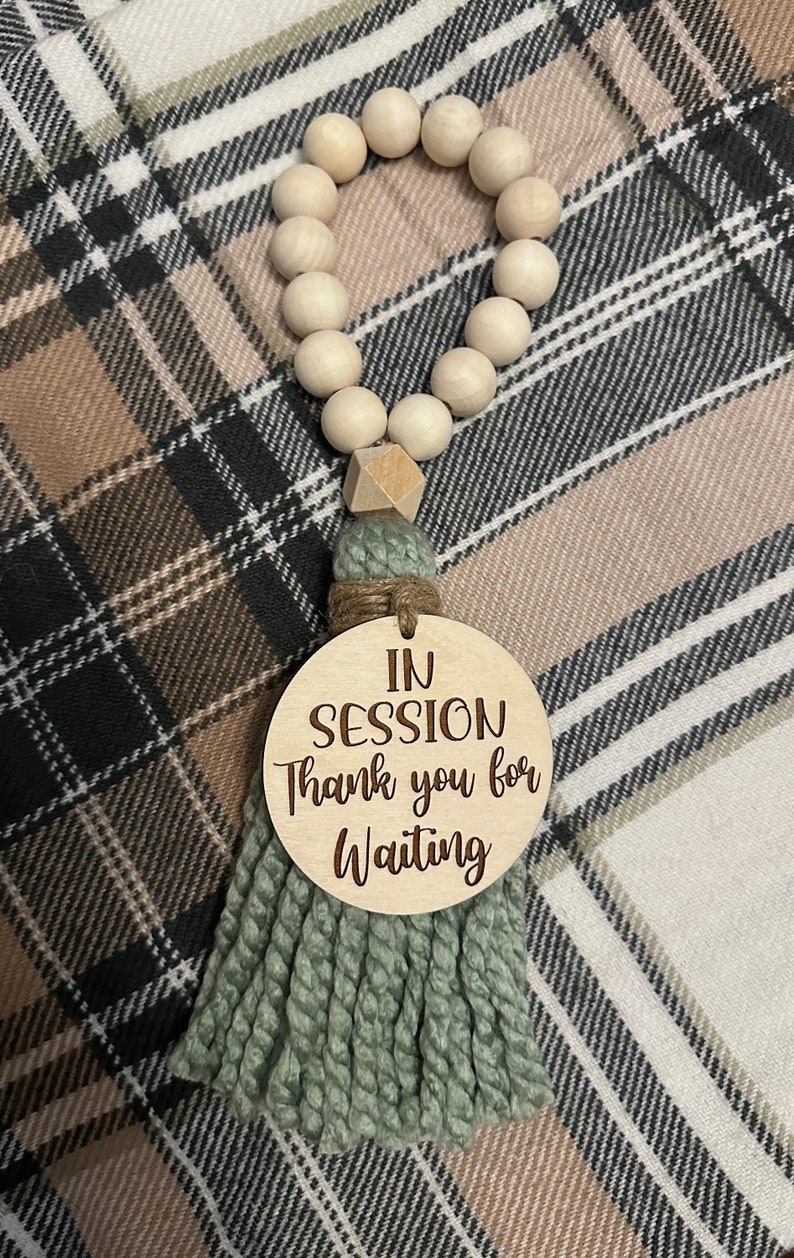 May include: A wooden bead garland with a sage green tassel and a round wooden sign that reads "IN SESSION Thank you for Waiting". The garland is made of light-colored wooden beads.